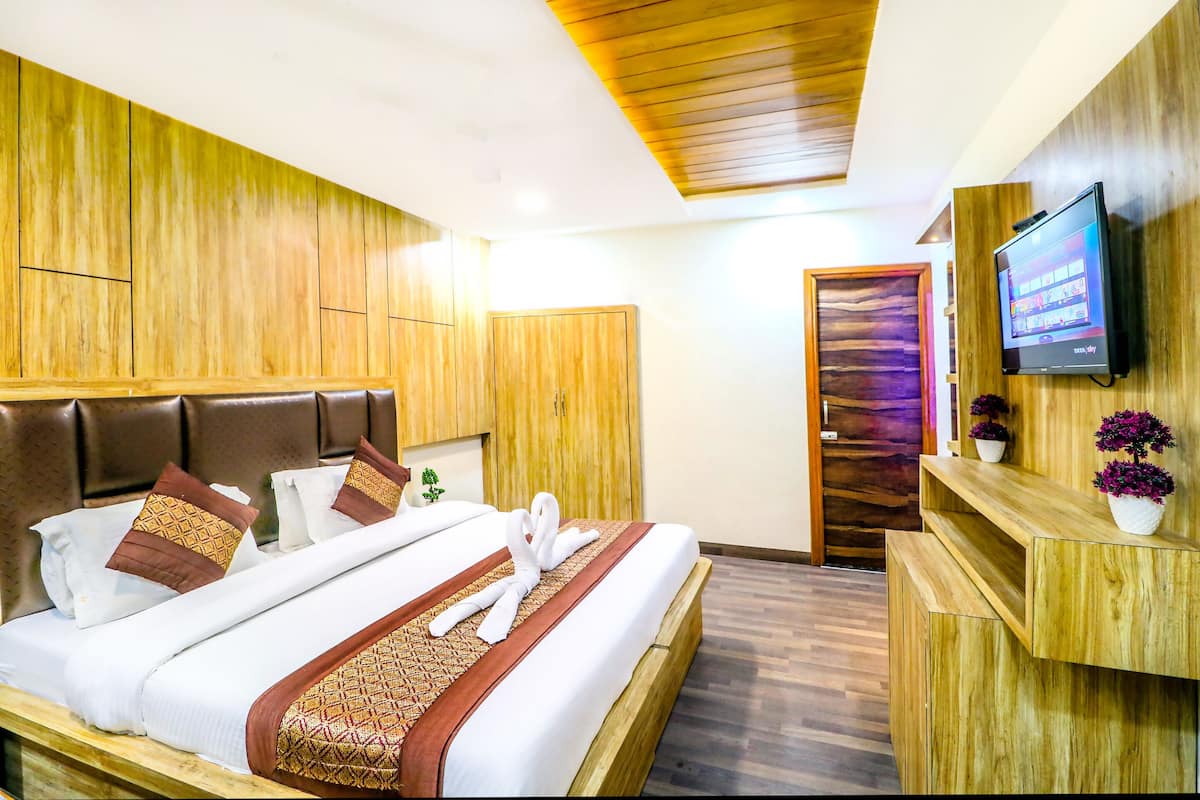 A hotel room with a wooden headboard, a bed with white bedding and decorative pillows, a wooden desk with a television, and a small plant.