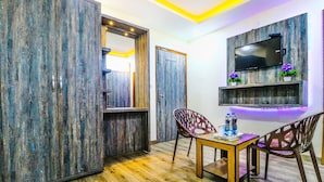 A room with a wooden wardrobe, a TV mounted on the wall, a dining table with chairs, and a small shelf with decorative plants. - Hotel City Castle Prime (Amritsar)