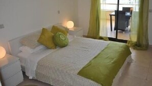 Ein Schlafzimmer mit Bett, Kissen, einem Nachttisch und Blick auf einen Außenbereich durch eine Glastür.