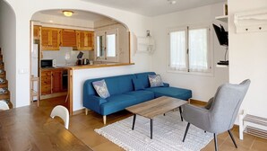 A compact living space with a blue sofa, a wooden coffee table, and a grey armchair. A ceiling fan is visible, and there is a kitchen area with wooden cabinets and a microwave. - 3 Bedroom Villa, private pool,  stunning Sea Views 5 mins from beach. WIFI & AC (Arenal D'en Castell)