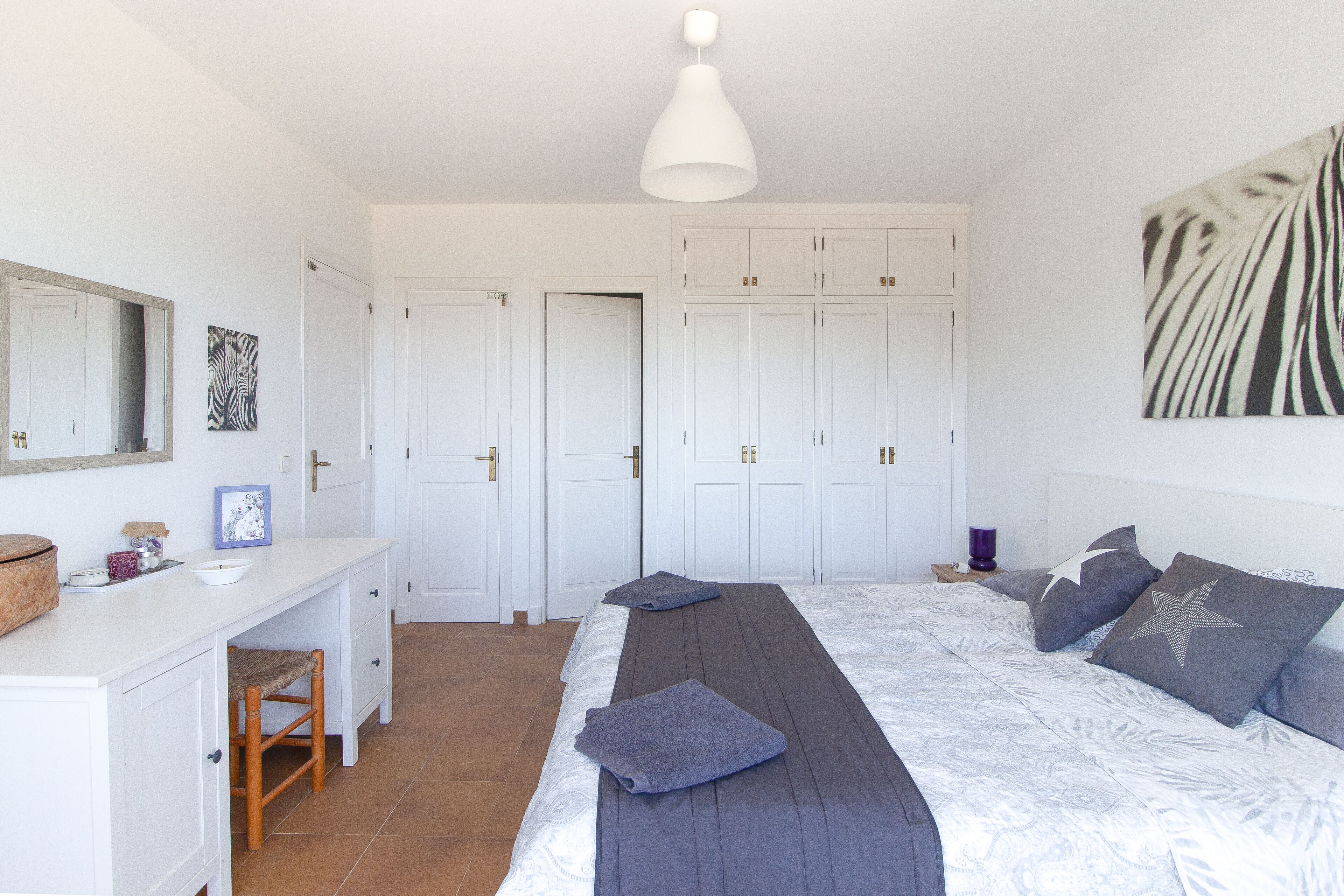A bedroom with a bed, a desk, and a chair. There are white wardrobes and a mirror on the wall.