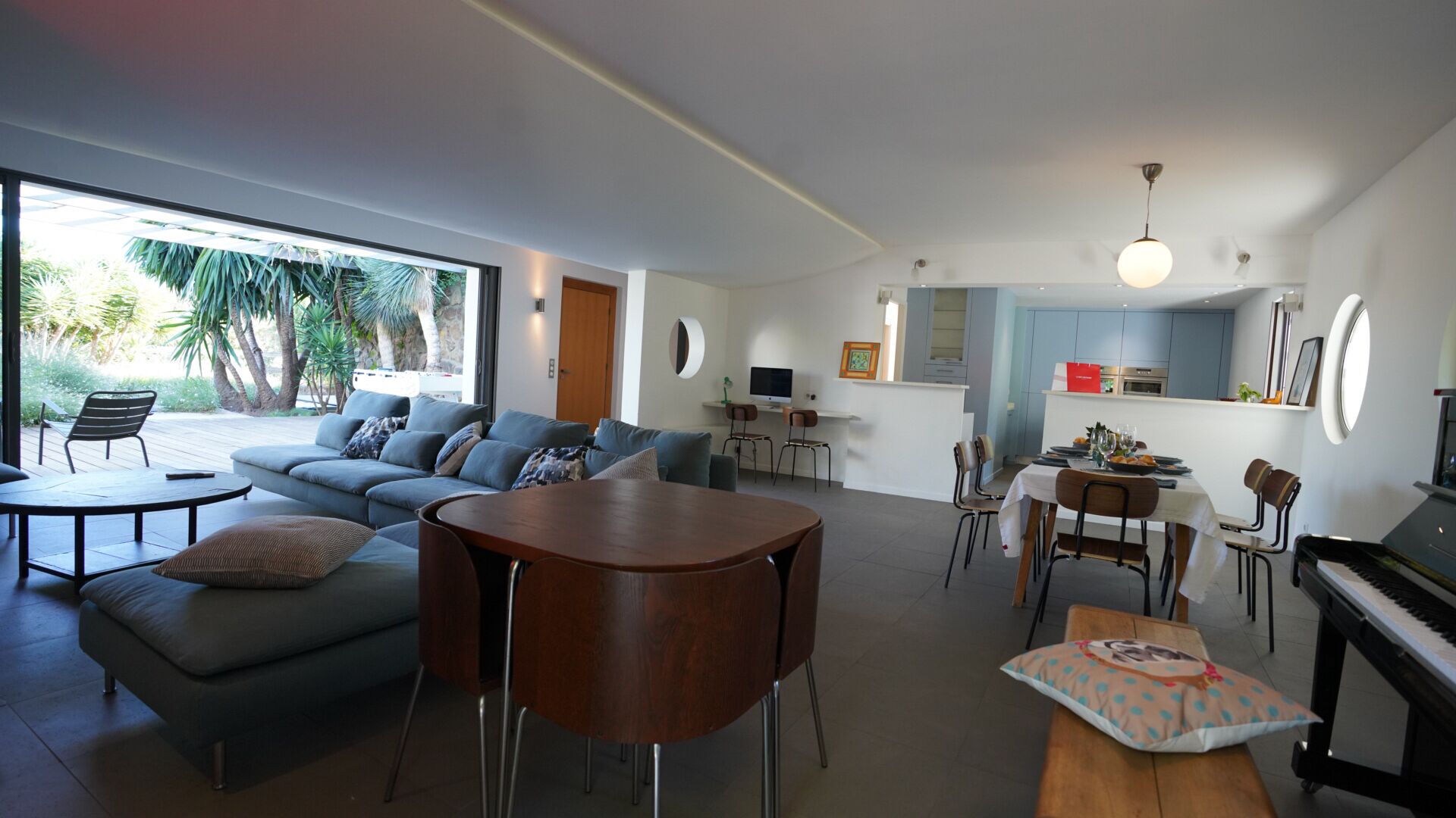 A modern living room with a sofa, a round wooden table, a dining area with chairs, a piano, and a kitchen area with cabinets and a countertop.