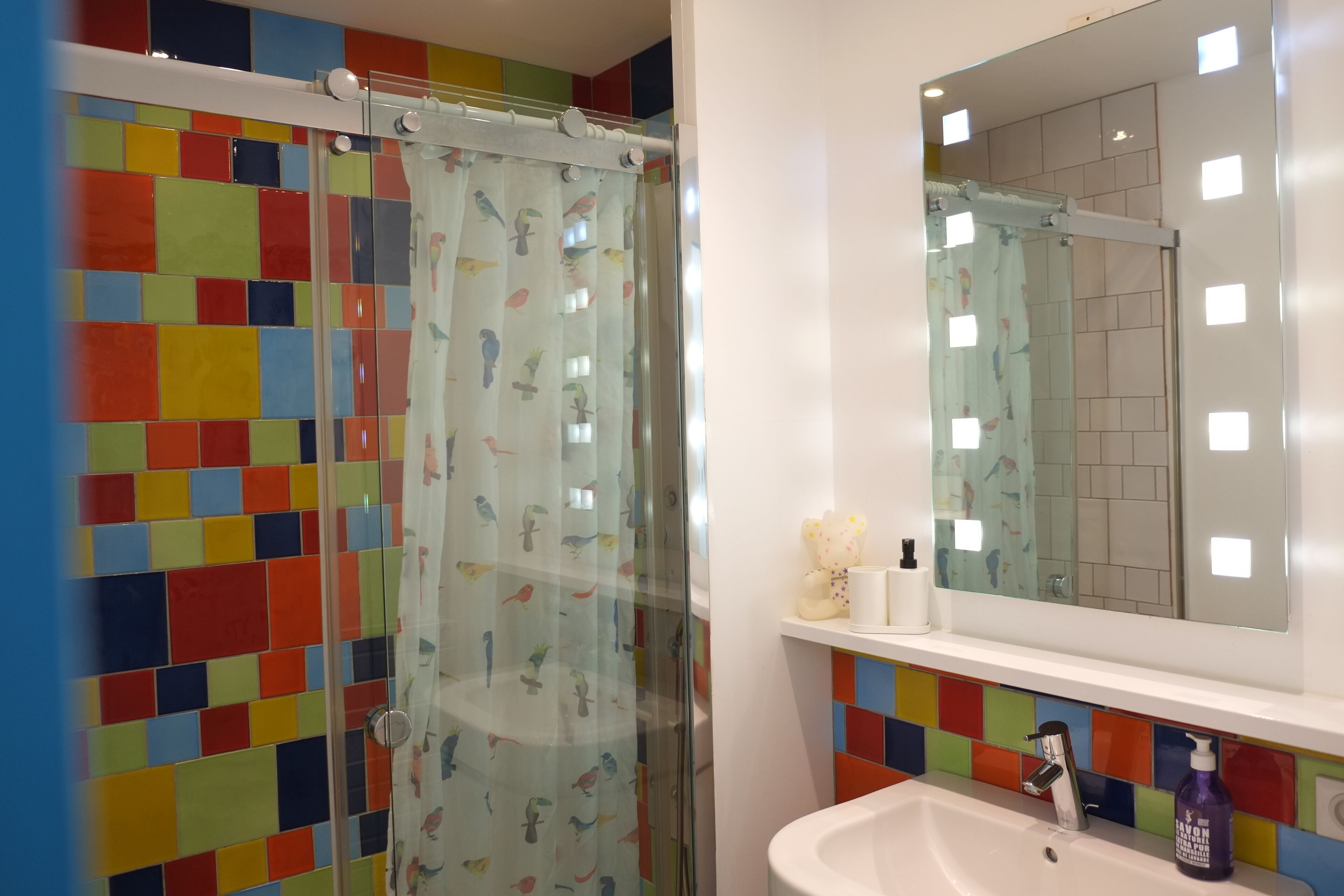 Een badkamer met kleurrijke tegels, een douchecabine van glas, een wastafel met zeepdispenser en een spiegel erboven.