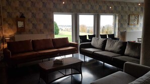 A living room with two sofas, a coffee table, and a radiator. There are large windows with a view of the outdoors. - Spacious and comfortable gîte with garden and very nice view  (Nassogne)