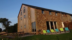 A stone building with a sloped roof and large windows. There are outdoor chairs and a bench near the entrance. - Spacious and comfortable gîte with garden and very nice view  (Nassogne)