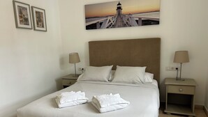 Un lit bien fait, avec du linge de lit blanc et deux oreillers, une tête de lit en bois, deux tables de chevet avec des lampes et, au mur, un tableau représentant un phare.