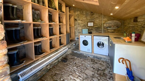 Une buanderie avec un sol en pierre, des étagères en bois où sont rangées des bottes, et deux machines à laver.