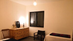 Un dormitorio con cama, cómoda, silla, ventana y mesita de noche con una botella.