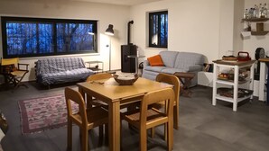 Una sala de estar acogedora con un sofá, una mesa de comedor con sillas, una chimenea y ventanas grandes con vistas a árboles.