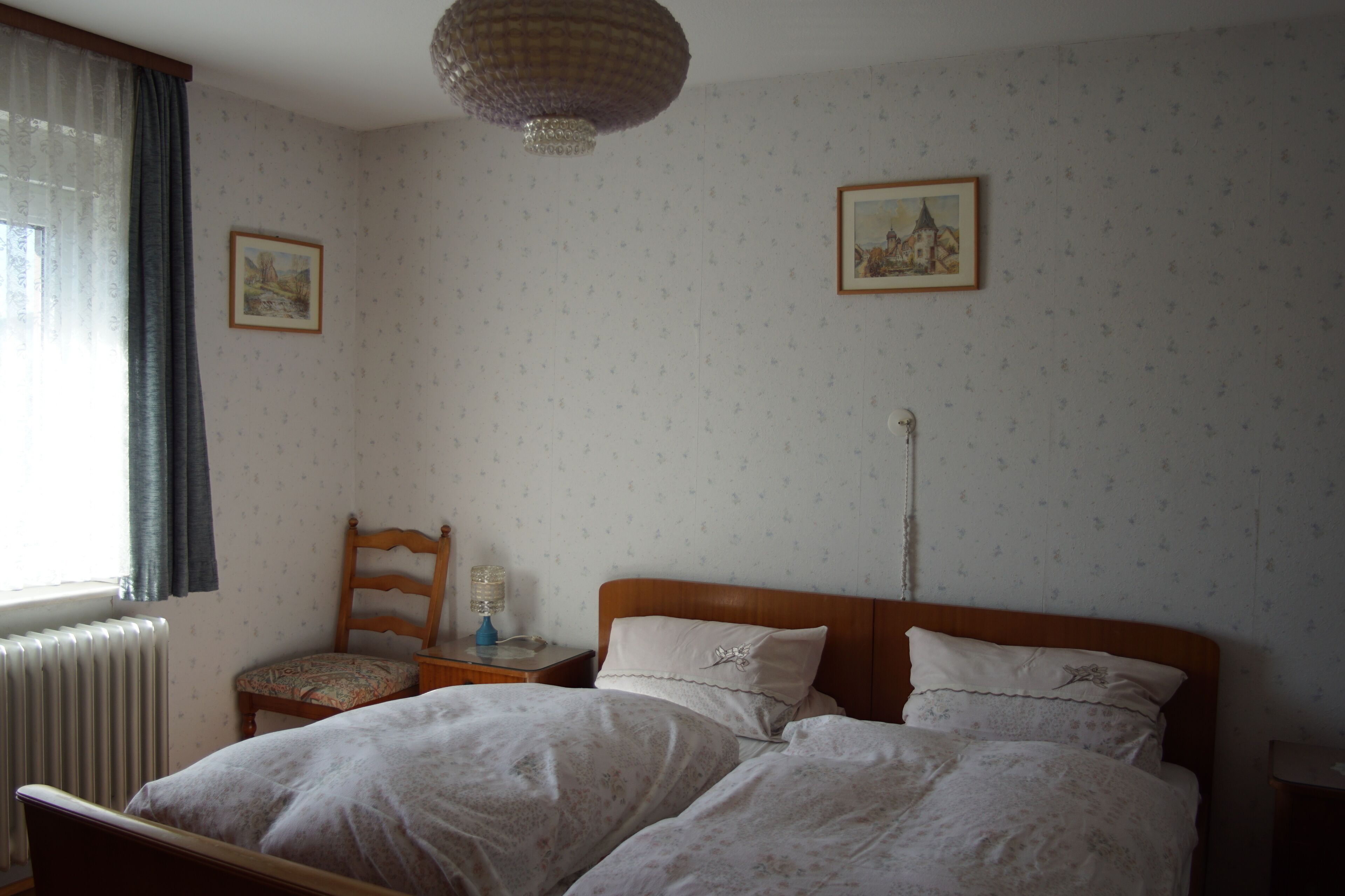 A bedroom with two beds, a chair, a nightstand, a radiator, and two framed pictures on the wall.