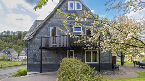 Een huis met twee verdiepingen en een balkon, omringd door groen en een heldere lucht.
