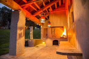 An outdoor stone fireplace with a fire, under a wooden roof structure, surrounded by stone walls and a grassy area. - Fürstenpalais holiday apartment in the tower of Schedling Castle near Lake Chiemsee (Trostberg)