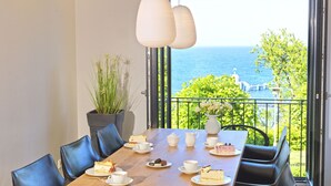 Una mesa de comedor con sillas, platos de comida, tazas y vista al mar a través de la barandilla de un balcón.