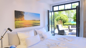 Habitación de hotel con una cama grande, un cuadro de un paisaje al atardecer, una lámpara de lectura y vistas del exterior a través de una puerta abierta.