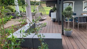 Terraza de madera con una mecedora colgante, una mesa y plantas en macetas.