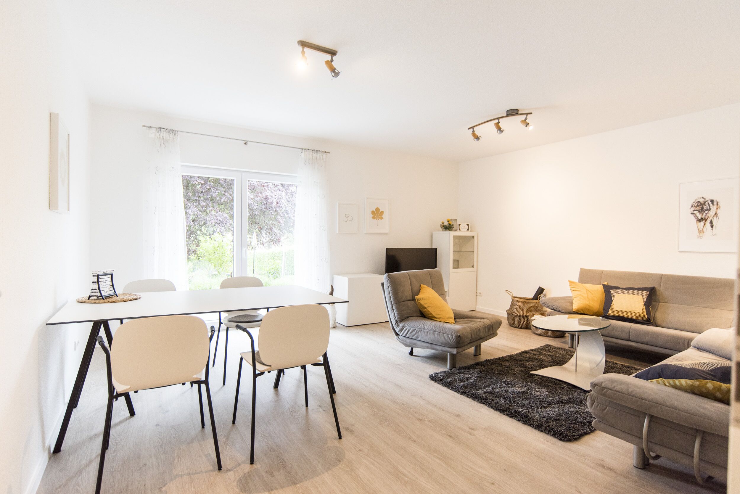 A modern living room with a dining area, a grey sofa with yellow cushions, a flat-screen TV, and a large window with sheer curtains.