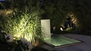 Una piscina moderna al aire libre con cerramiento de cristal, rodeada de exuberante follaje y una colina boscosa oscura al fondo.