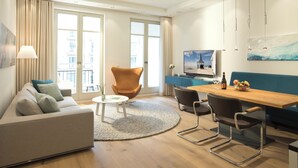 A modern living room with a sofa, a round coffee table, a dining table with chairs, a TV, and a view of the city through large windows. - Summerbreeze Cloud Luxury Apartment with Ocean View | Skypool, Sauna & Beach Chair | Dog-Friendly (Sellin)