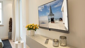 A modern living room with a large flat-screen TV mounted on the wall, a vase with yellow flowers on a console table, a remote control, and two decorative glass vases. - Summerbreeze Cloud Luxury Apartment with Ocean View | Skypool, Sauna & Beach Chair | Dog-Friendly (Sellin)
