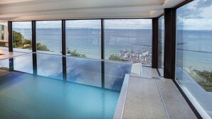 A modern infinity pool with a clear view of the sea and a coastal town. - Summerbreeze Cloud Luxury Apartment with Ocean View | Skypool, Sauna & Beach Chair | Dog-Friendly (Sellin)