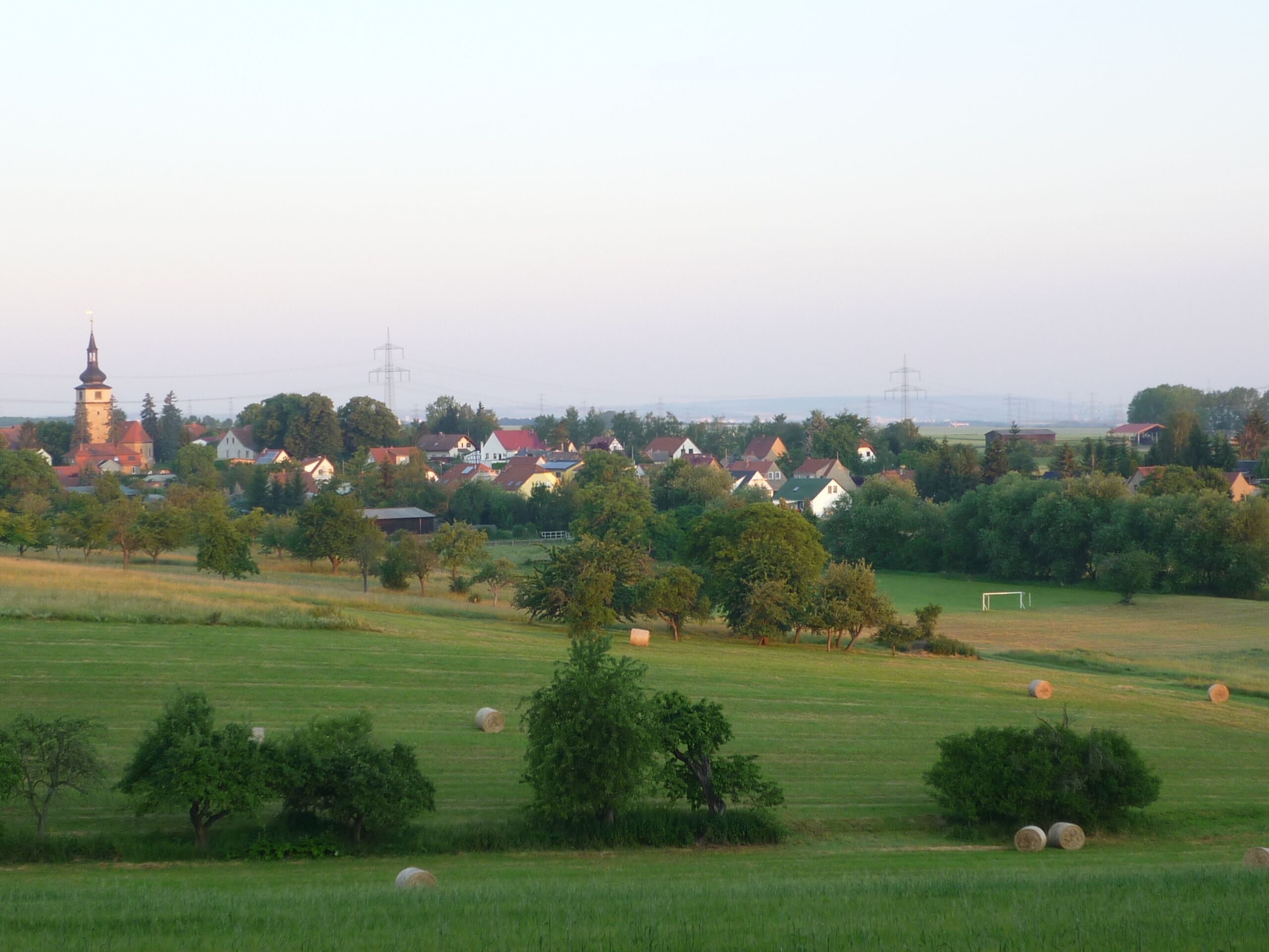 Un paysage rural avec des maisons, un clocher d’église et des bottes de foin dans un champ.