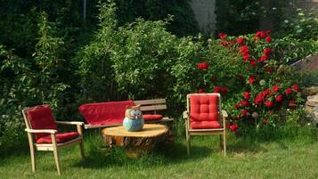 A garden with two red chairs and a wooden table, surrounded by lush greenery and red flowers.