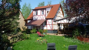 Uma casa tradicional de enxaimel com telhado inclinado, um jardim com relvado, duas cadeiras de jardim e um banco de madeira.