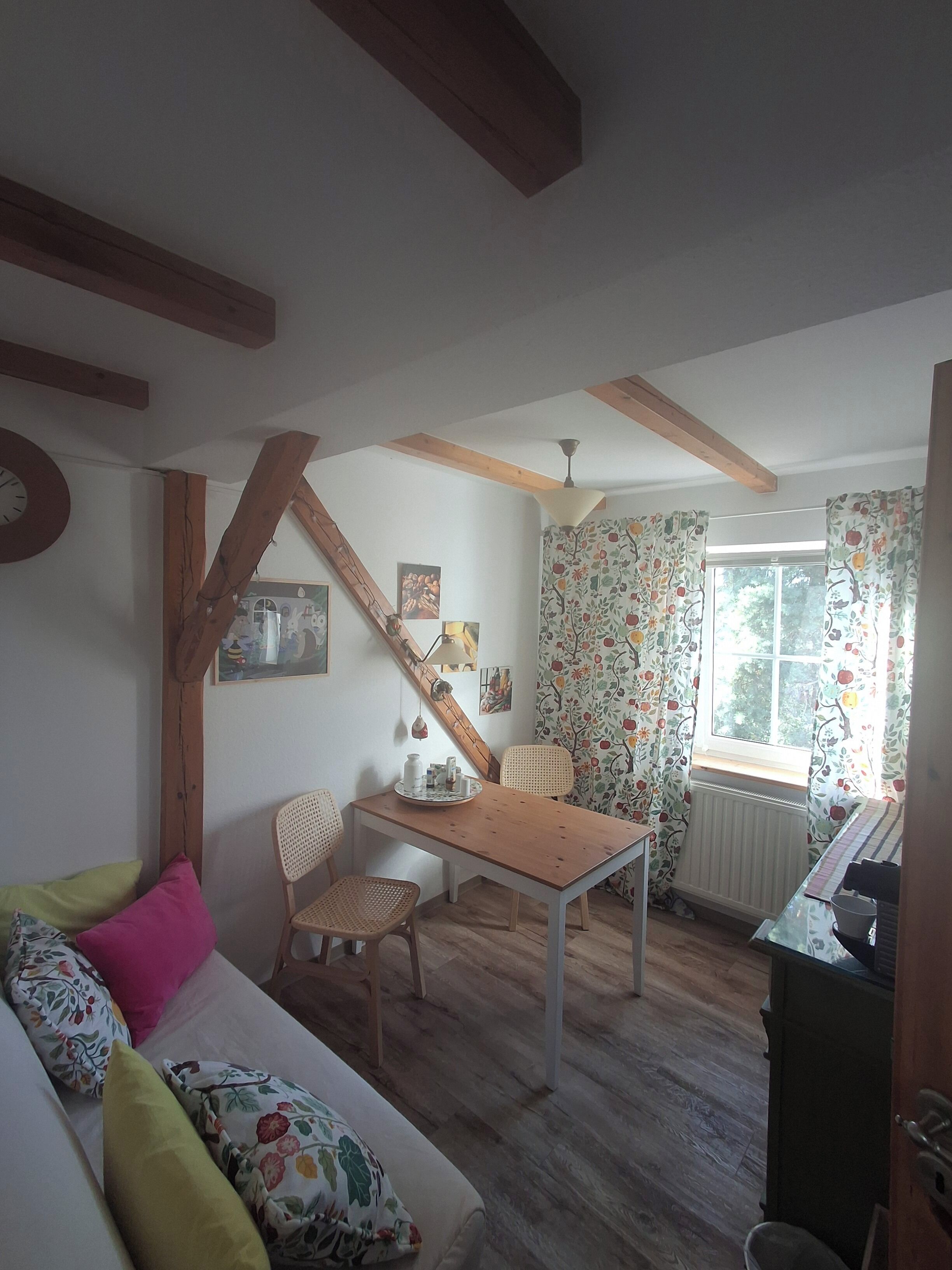 Une pièce chaleureuse avec une table en bois, un canapé agrémenté de coussins à motifs floraux, une fenêtre avec des rideaux et un radiateur.