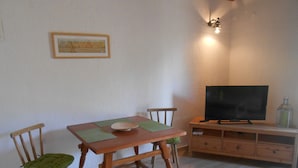Zona pranzo con tavolo in legno, due sedie, un televisore su un mobile basso e un piccolo armadietto.
