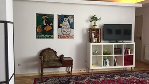A living room with a wooden chair, a white TV stand with books, a red patterned rug, and two posters on the wall. - Apartment in the heart of Kreuzberg, loft,art,pubs,bars,nightlife,WiFi,up to 4 persons  (Bezirk Friedrichshain-Kreuzberg)