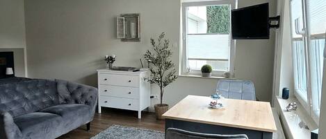 Een moderne woonkamer met een grijze bank, een eettafel met stoelen, een kleine potplant en een raam met jaloezieën.