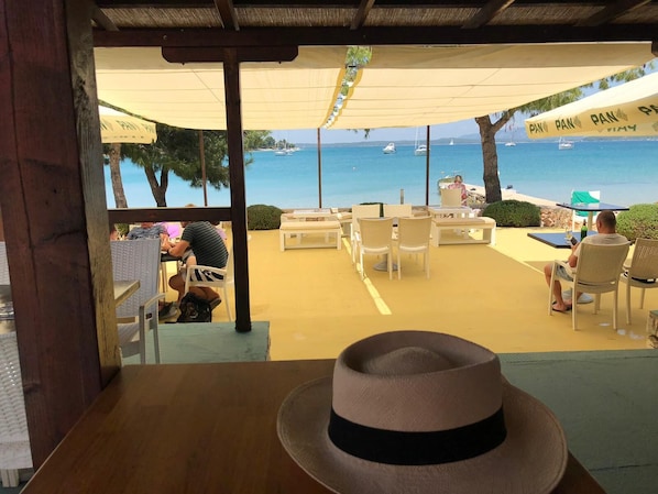 An outdoor seating area with tables and chairs, overlooking a beach with boats.