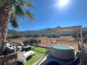 Una terraza de madera con un jacuzzi, sillones de descanso y una pérgola con vistas a un paisaje montañoso.