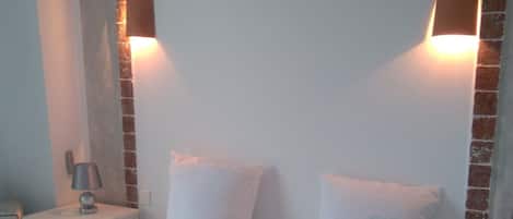Una cama bien hecha con sábanas y almohadas blancas, dos mesitas de noche con lámparas y una pared decorativa de ladrillo.
