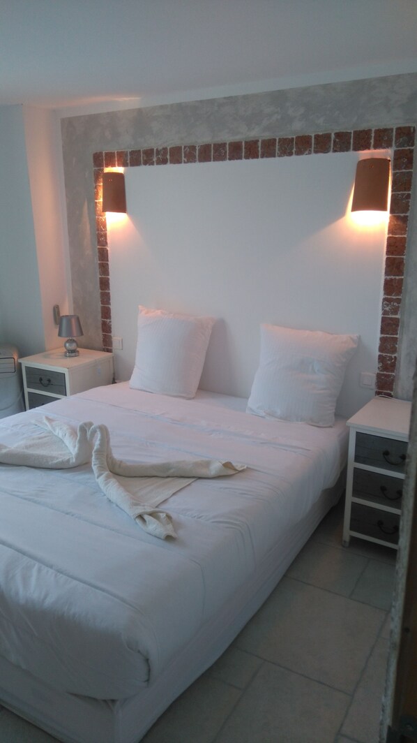Una cama bien hecha con sábanas y almohadas blancas, dos mesitas de noche con lámparas y una pared decorativa de ladrillo.