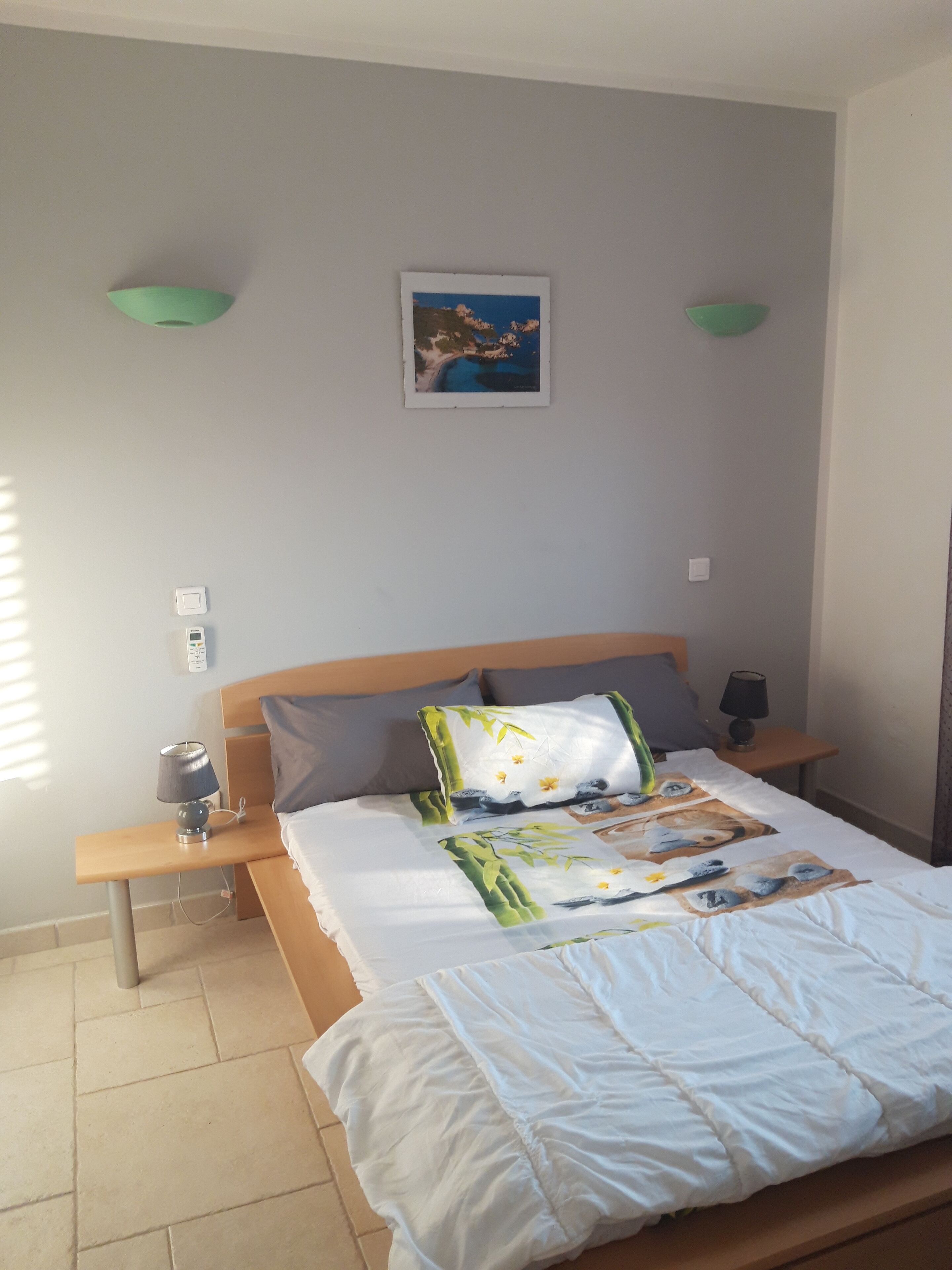 Una cama bien hecha con cubrecama blanco y manta estampada con flores, cabecera de madera, dos mesitas de noche con lámparas y un cuadro colgado en la pared que muestra una escena costera.