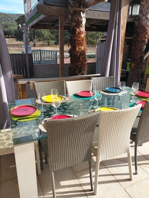 Un área de comedor al aire libre con sillas de mimbre y una mesa de vidrio, frente a un telón de fondo de palmeras y vistas al campo.