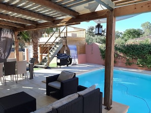 Área externa coberta com piscina, móveis de vime, churrasqueira e escadaria que dá para um deck de madeira.
