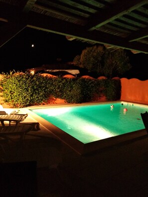 Una piscina al aire libre de noche con algunas personas nadando, rodeada de vegetación y unas cuantas sillas de descanso.