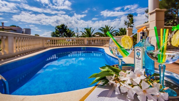 En pool med krystalklart, blåt vand, omgivet af en hvid gelænder, med et bord i forgrunden, hvor der står en flaske Bombay Sapphire Gin, et glas med et citronskive og et grønt sugerør, samt hvide blomster.