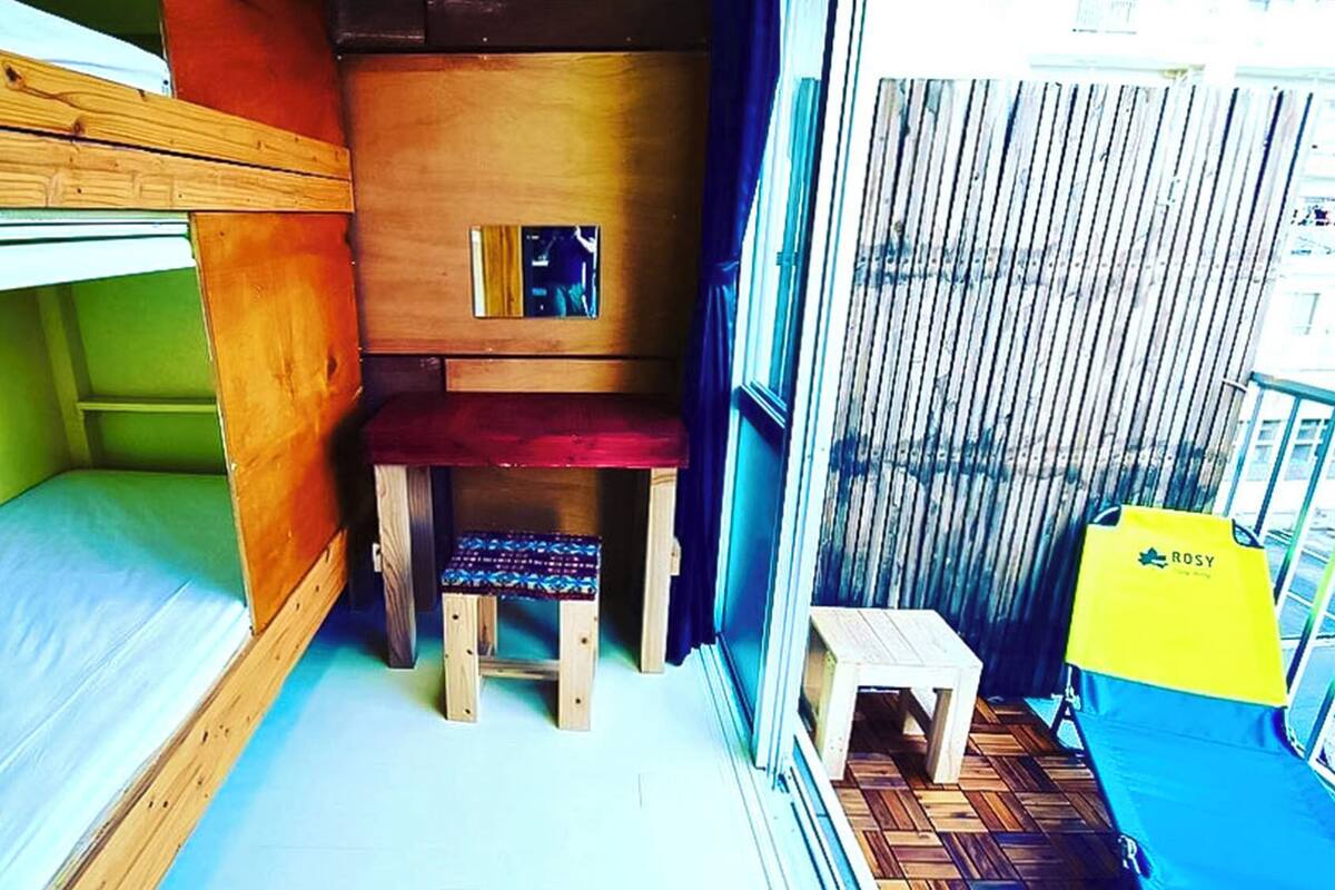 Une petite pièce comprenant un lit superposé, une petite chaise en bois, une petite table et un sac jaune.