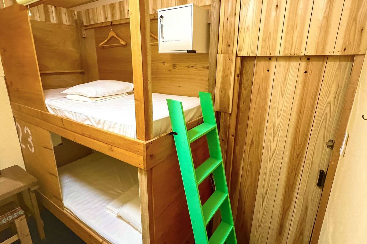 Une chambre avec un lit superposé en bois, une échelle verte, un petit bureau et un luminaire suspendu.