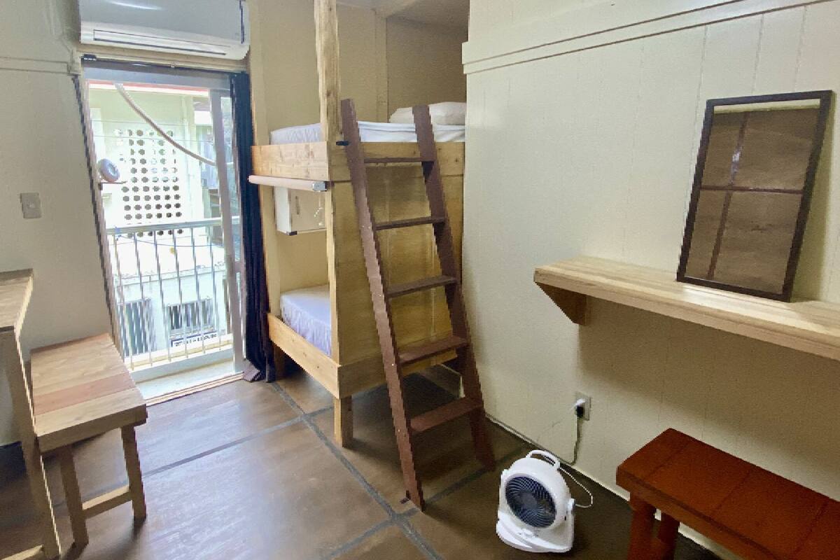 Une petite pièce, de taille modeste, comprenant un lit superposé, un bureau en bois, une petite table, une échelle et un ventilateur.