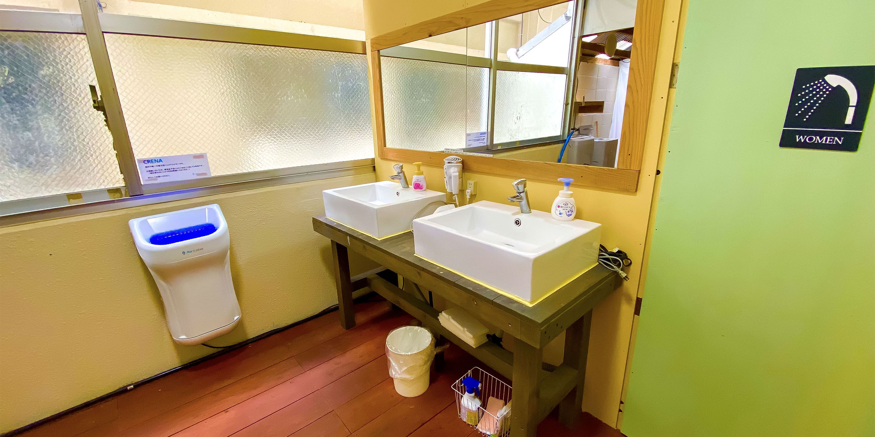 A women's restroom with two sinks, a mirror, a hand dryer, and a trash bin.