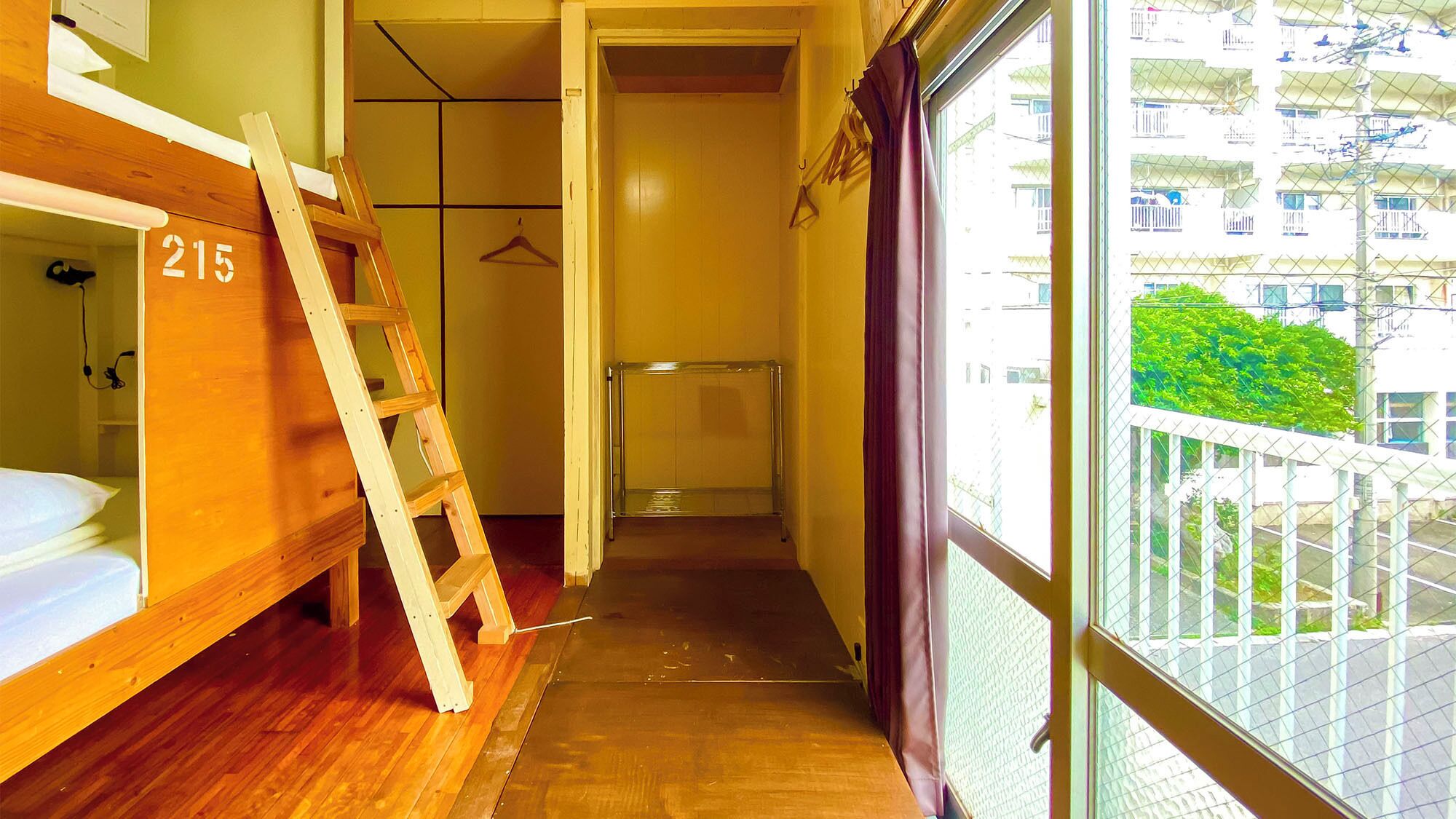 Un pasillo estrecho con una cama litera de madera con el letrero '215', un estante de vidrio transparente y vistas a un balcón con vegetación.