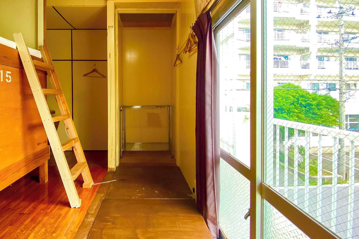 Un couloir étroit avec un lit superposé en bois portant le numéro « 215 », une étagère en verre transparent et une vue sur un balcon agrémenté de plantes.