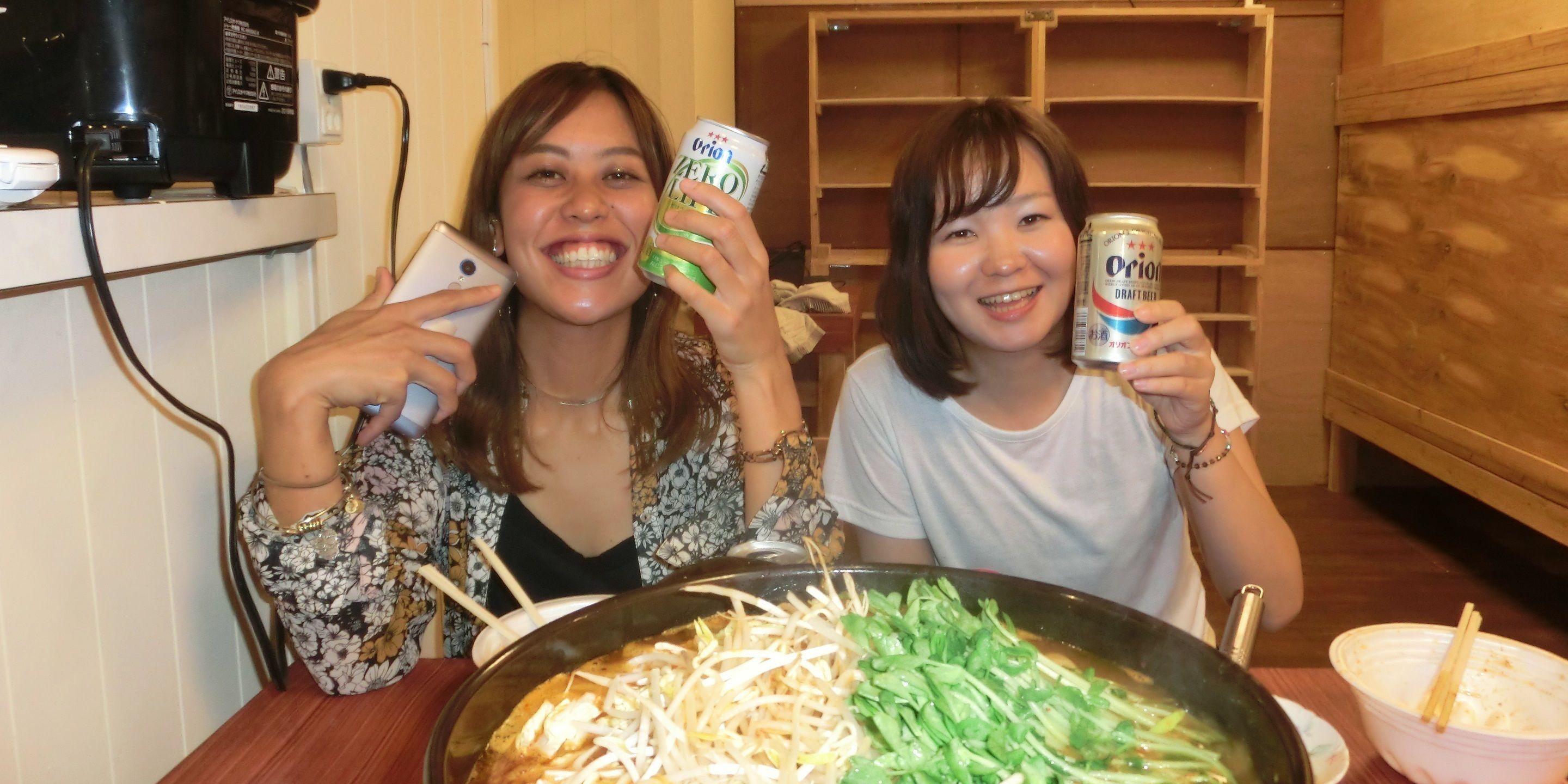 Twee personen houden blikjes drinken vast, zittend aan een tafel met een grote schaal eten ervoor.
