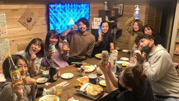 Un grupo de personas alrededor de una mesa de madera con comida y bebidas, un televisor al fondo y paredes de madera.
