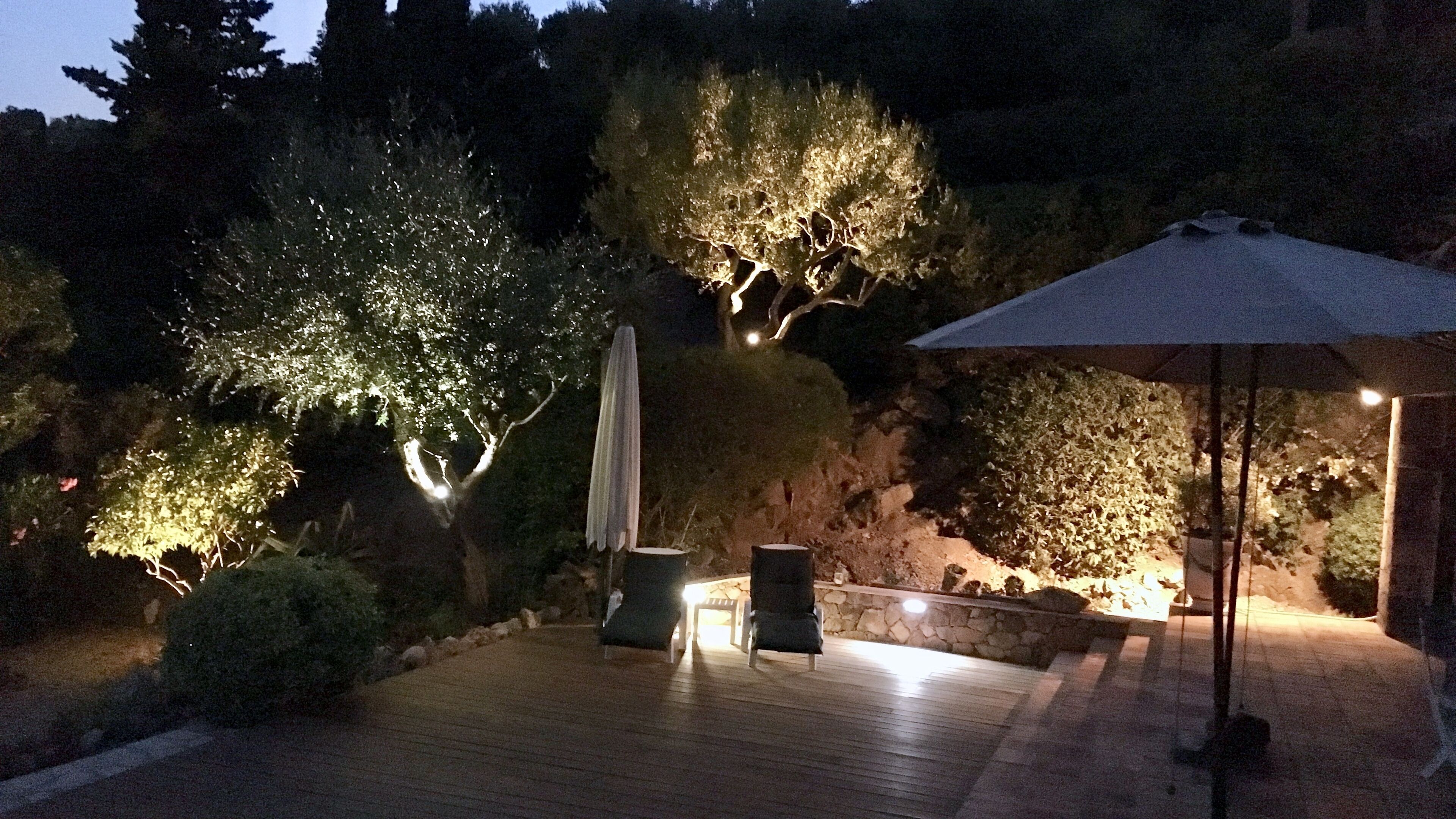 A well-lit outdoor patio area with wooden flooring, two chairs, a table, and an umbrella.