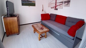 Ein Wohnzimmer mit grauem Sofa, roten Kissen, einem Holztisch, einem Fernseher auf einer Holzkommode und Wandbildern, darunter eine Brücke und ein Schloss.
